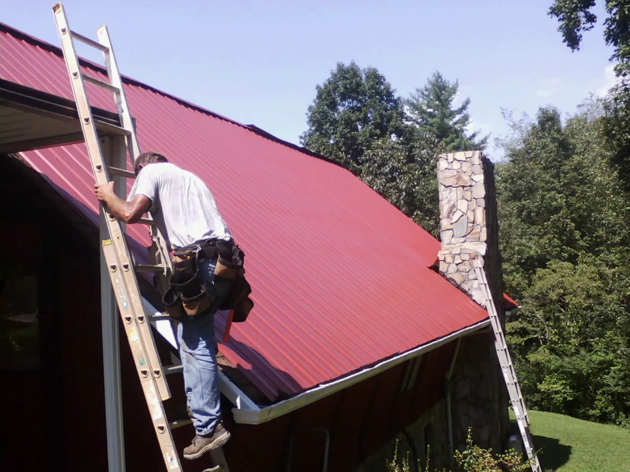 Roofer on a ladder beside a red metal roof and stone chimney — Soffit & Fascia Repair by Roofing Brothers in Salmon